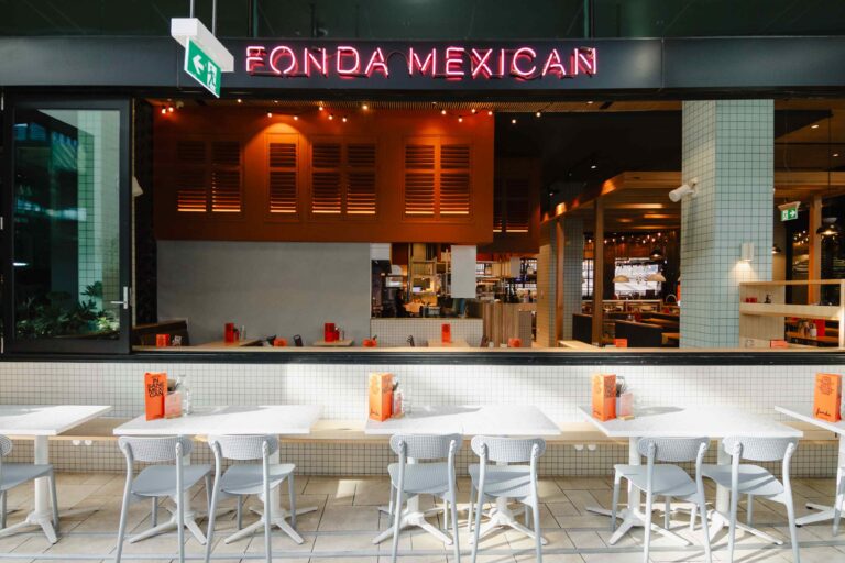 A view of the storefront of a restaurant named Fonda Mexican. In the foreground is an outdoor seating area with row of white tables and light grey chairs set up against a low wall.