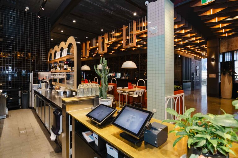 A front view of the interior of a modern restaurant and bar fitout for Fonda Mexican Chadstone. The space features a long, stainless steel counter with a bar area on the left, displaying bottles and glasses.