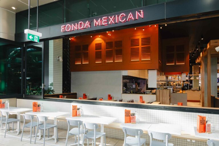 A view of the storefront of a restaurant named Fonda Mexican. In the foreground is an outdoor seating area with row of white tables and light grey chairs set up against a low wall.