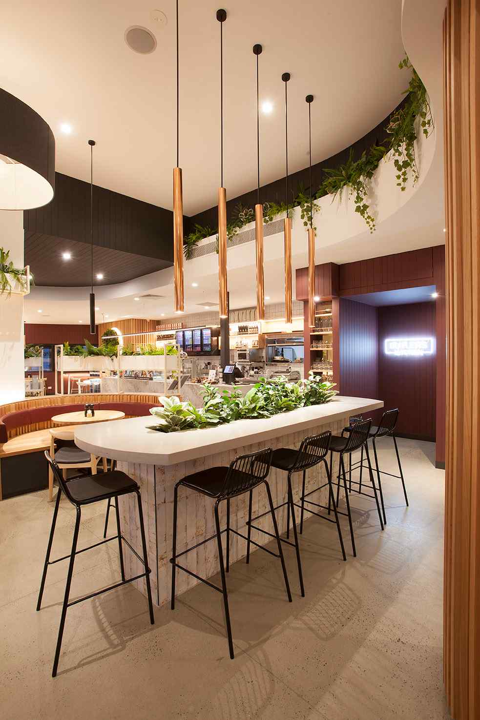 A café communal seating area featuring a large central counter with high black stools. The countertop is decorated with lush green plants, and copper pendant lights hang from the ceiling. In the background, curved booth seating and the service counter are visible.