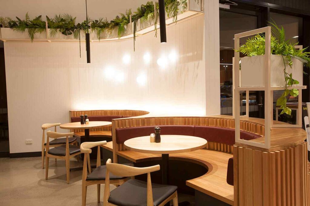 A cozy café seating area with curved wooden booths and round tables. The booths are upholstered with burgundy backs, paired with light wooden chairs and black cushions. Above, a shelf with cascading green plants adds freshness to the warm-toned space.